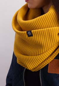 Yellow knitted cowl neck scarf featuring a ribbed texture and a small black logo tag, paired with a dark quilted jacket.