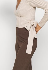 Beżowy sweter z prążkowanej dzianiny z długim rękawem i zawiązaniem z tyłu, zestawiony z ciemno-brązowymi spodniami o wysokiej talii i prążkowanej strukturze.