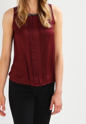 Ciemnoczerwona bluzka na ramiączkach z pionowymi plisami na przodzie oraz czarne spodnie, noszone przez stojącą osobę o rozpuszczonych blond włosach.