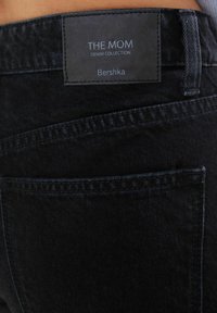 Svart denimjeans med en etikett som säger "THE MOM DENIM COLLECTION" av Bershka. Har subtil struktur och standard fickdesign.