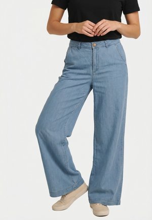 Mujer con pantalones vaqueros anchos de pierna ancha azul claro, camiseta negra de manga corta y zapatillas deportivas beige, de pie con las manos entrelazadas al frente.