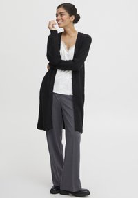 Schwarzer, lang geschnittener Cardigan mit weicher Textur, offene Vorderseite, kombiniert mit einem weißen Oberteil mit Spitzenbesatz und grauen, weit geschnittenen Hosen, getragen mit schwarzen Schuhen.