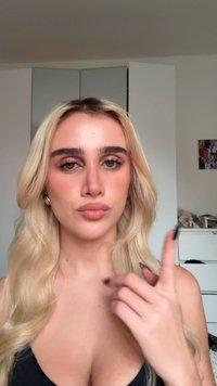 En blond kvinne med langt hår og sminke peker oppover med høyre hånd i et rom med hvite skap og et collage på veggen.