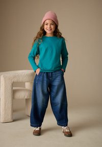 Türkisfarbenes Langarmshirt, lockere blaue Jeanshose mit umgeschlagenem Saum und braune Schuhe mit Leopardenmuster. Das Modell trägt eine rosa gestrickte Mütze.