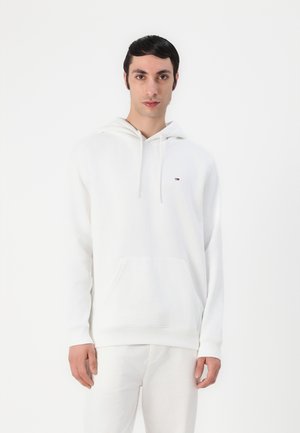 Sudadera blanca de algodón con un bolsillo canguro en la parte frontal, capucha con cordón y un pequeño logotipo en el lado izquierdo del pecho. La tela parece suave y lisa.