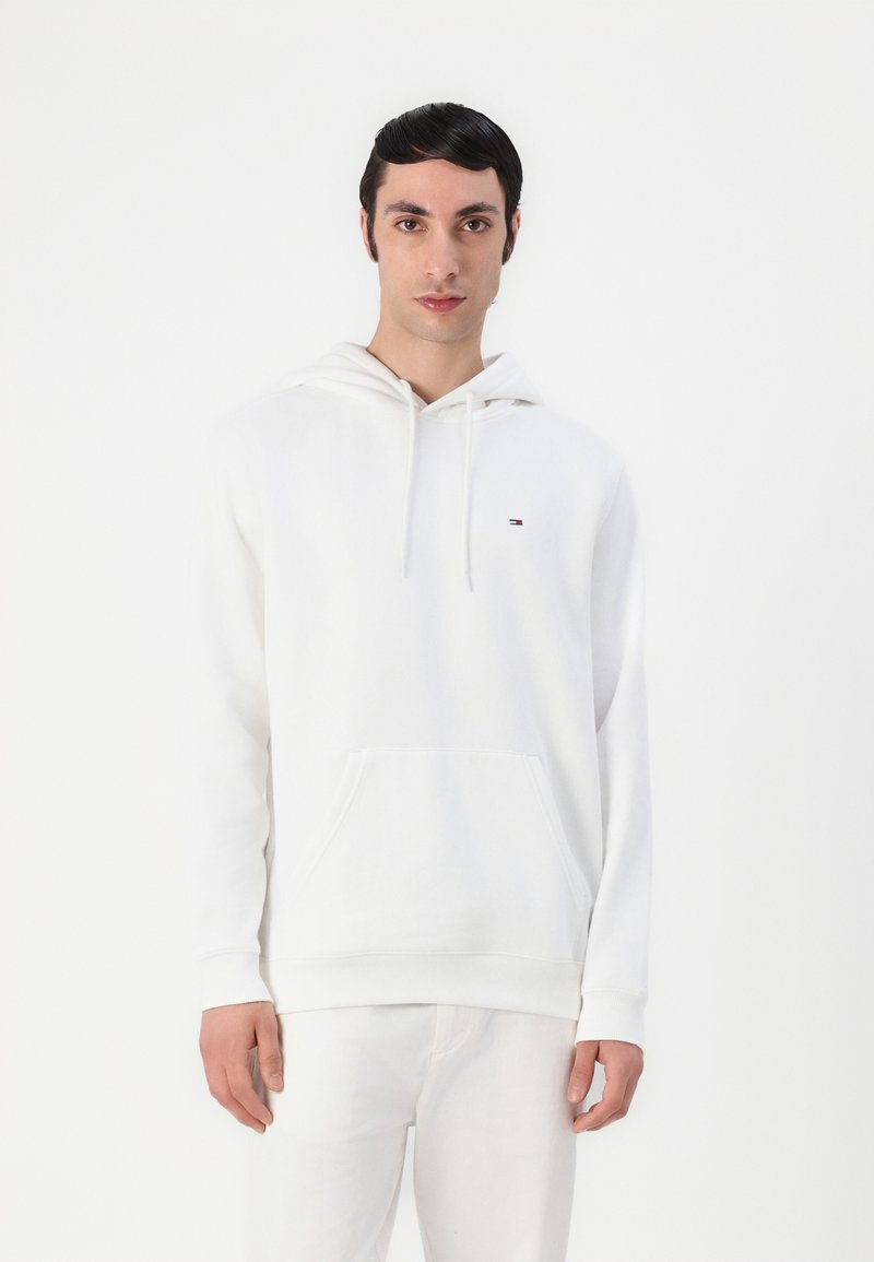 Hoodie en coton blanc avec une poche kangourou à l'avant, capuche à cordon et un petit logo sur la poitrine gauche. Le tissu semble doux et lisse.