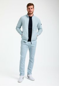 Giacca blu chiaro trapuntata con zip abbinata a pantaloni coordinati. Camicia blu scuro sotto. Sneakers bianche con dettagli neri. Texture liscia.