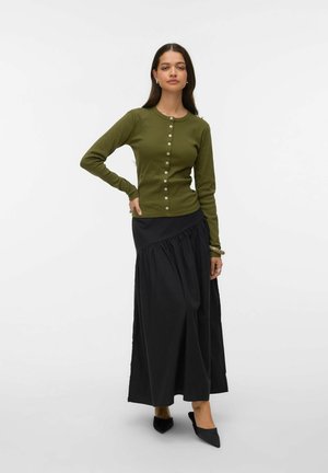 Žena stojící s rukou v bok, oblečená v olivově zeleném dlouhém tričku na knoflíky, černé asymetrické maxisukni a černých špičatých botách.
