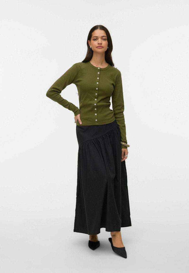 Žena stoji s rukom na bok, nosi olivno zelenu dugih rukava s dugmadima, crnu asimetričnu maksi suknju i crne šiljate cipele.