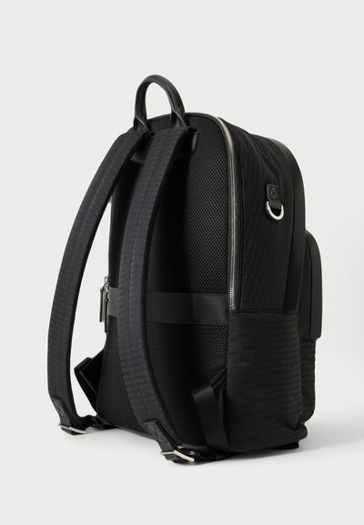 Sac à dos noir avec bretelles rembourrées et réglables, panneau dorsal en mesh, compartiment principal zippé et anneau en D en métal sur le côté.
