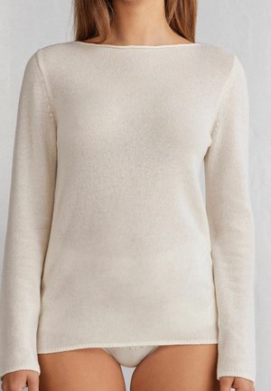 Donna che indossa un maglione a maniche lunghe aderente color crema, lavorato a maglia, con scollo a barca, corto appena sopra i fianchi.
