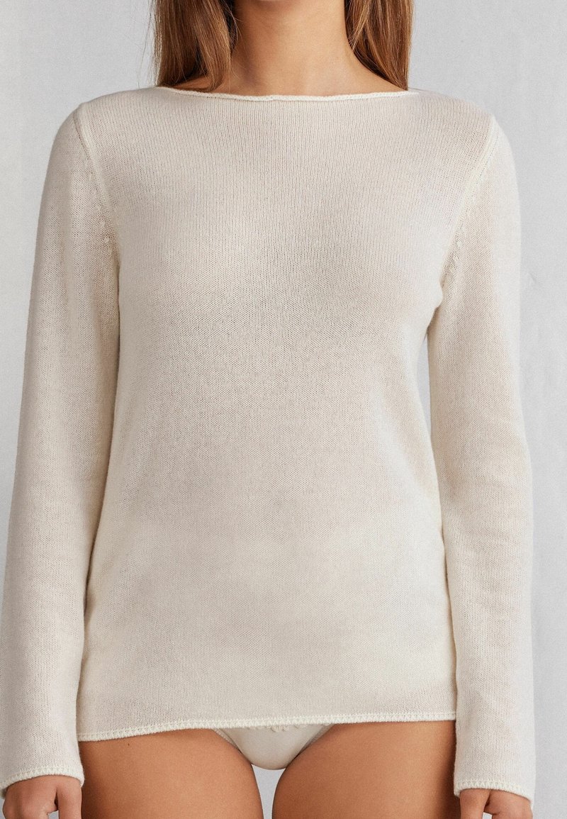Femme portant un pull ajusté en maille crème à manches longues avec un col bateau, coupé juste au-dessus des hanches.