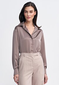 Jasnobrązowa satynowa bluzka z ostrym kołnierzykiem i guzikiem z przodu, w zestawieniu z jasnobeżowymi spodniami w prążki, podkreślającymi gładkie faktury.