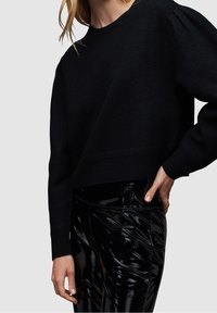 Schwarzer, kurzer Pullover mit langen Ärmeln und Rundhalsausschnitt, aus strukturiertem Stoff gefertigt. Kombiniert mit glänzenden schwarzen Leggings mit glatter Oberfläche.