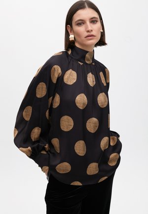 Woman wearing a black blouse with large textured gold polka dots, high neck, puffed sleeves, and gold statement earrings against a white background.