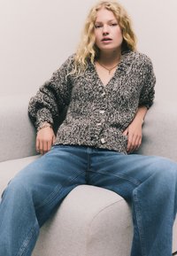 Rūtaini brūns un melns tamborēts kardigans ar V-veida izgriezumu, lielām pogām un ribotiem aprocēm, valkāts kopā ar gaiši zilām plaša piegriezuma džinsu biksēm.