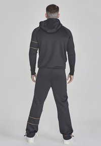 Survêtement sportif noir en matière douce, comprenant une veste à capuche avec des bandes dorées et un pantalon assorti avec des motifs aux chevilles.