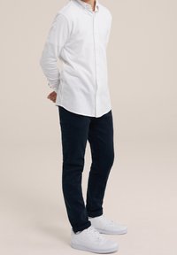 Person klädd i en vit skjorta med knappar, mörka byxor och vita sneakers, stående med händerna bakom ryggen på en neutral bakgrund.