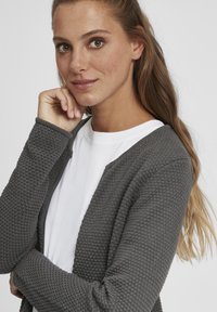 Gray textured cardigan made of knitted fabric, featuring a relaxed fit and open front design. Worn over a white shirt, showcasing a casual style.