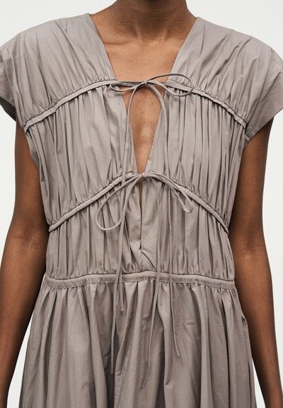 Žena v taupe bezrukávových šatách so zhrnutou látkou a dvoma viazanými mašľami vpredu na výstrihu a v oblasti pása.