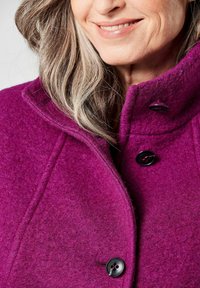 Fuchsia Wollemantel mit hohem Kragen, ausgestattet mit schwarzen Knöpfen und einer strukturierten Oberfläche. Nahaufnahme des Kragens und der Knopfdetails.