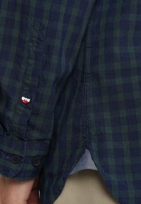 Rutig skjorta med knappar i marinblått och grönt tyg. Har två svarta knappar på manschetten och en liten tri-färgad detalj vid knappen.