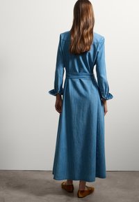 Abito maxi in denim con maniche lunghe, vita aderente e cintura annodata. Presenta una texture liscia e un orlo leggermente svasato, mostrato da dietro.