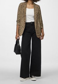 Žena v saka s leopardím potiskem, bílém topu, černých kalhotách s širokými nohavicemi, černých teniskách a držící malou černou kabelku.