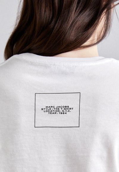 Hvit bomull t-skjorte med et rektangulært svart merkelapp på baksiden med tekst: "MARC JACOBS STIL: T-SKJORTEN STED: N.Y.C. ÅR: 1984."