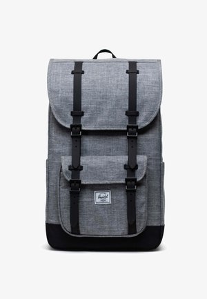 Sac à dos en tissu gris avec une fermeture à rabat, des bretelles noires, une poche avant et un patch logo. Matériau texturé avec un design bicolore.