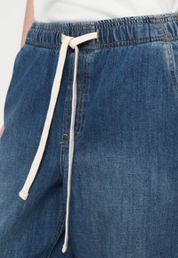 Μπλε τζιν παντελόνι με ελαστική μέση και λευκό κορδόνι δεμένο σε φιόγκο μπροστά, φορεμένο με λευκή μπλούζα.