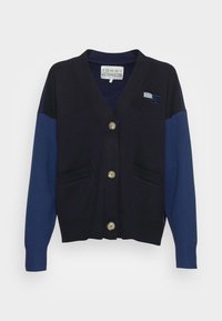 Cárdigan azul marino con mangas azules en contraste, textura acanalada y cuatro grandes botones dorados. Presenta dos bolsillos frontales y un acento con el logo.