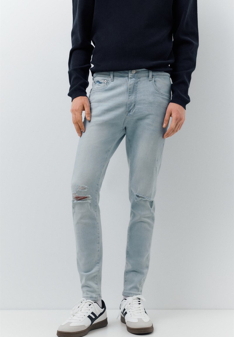 Homme portant un jean bleu clair déchiré, un chemisier noir à manches longues et des baskets blanches, debout devant un fond blanc uni.