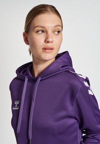 Felpa con cappuccio viola caratterizzata da un design elegante, chiusura con coulisse e dettagli bianchi sulle spalle, con un logo sul petto.