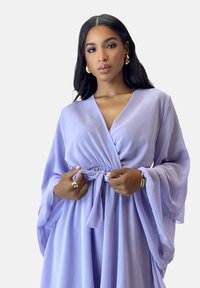 Vestito avvolgente color lavanda in tessuto leggero, caratterizzato da vita aderente, profonda scollatura a V e maniche a campana fluide. Texture liscia e morbida.