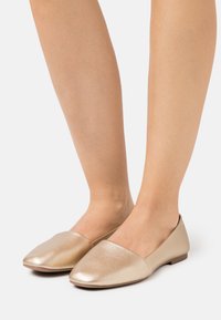 Litte ballet kengät kiiltävässä metallinhohtoisessa kultaisessa nahassa, joissa on terävä kärki ja minimalistinen muotoilu. Kengissä on matala korko ja yksinkertainen siluetti.