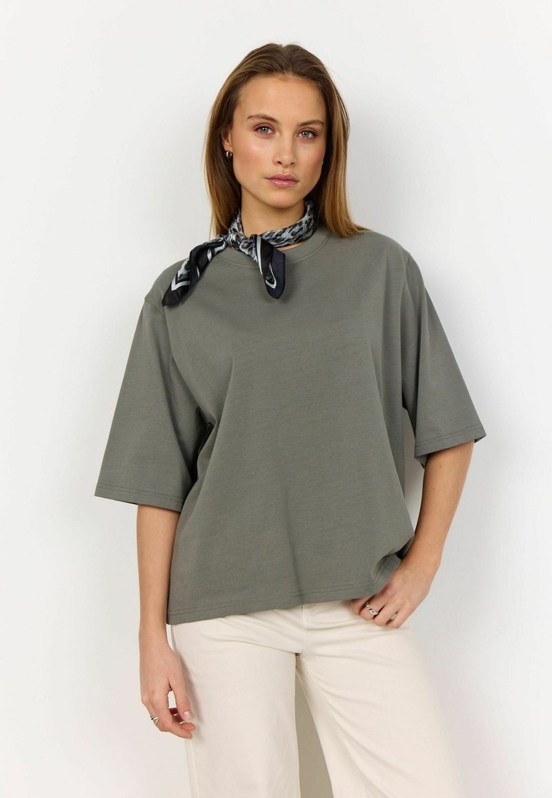 Olivenfarvet oversized t-shirt med korte ærmer, ensfarvet tekstur, parret med et sort og hvidt mønstret silkesjal bundet omkring halsen.