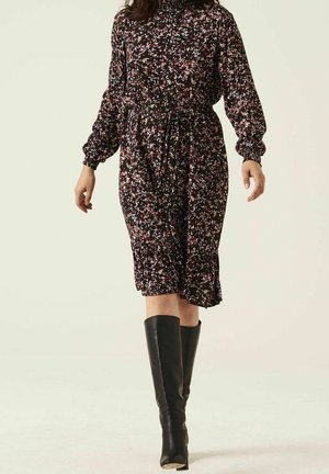 Vrouw met een lange mouwen, afgeronde bloemen midi-jurk met ceintuur en kniehoge zwarte leren laarzen, staand tegen een effen achtergrond.