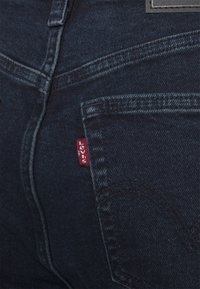 Gros plan sur un jean en denim bleu foncé montrant la poche arrière avec une étiquette en tissu rouge Levi's et des détails de couture visibles.