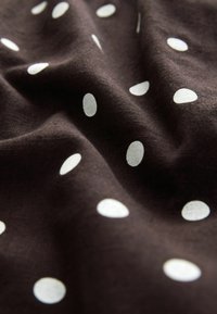 Ciemnobrązowa tkanina w białe, owalne kropki. Materiał wydaje się mieć fakturę i miękki spadek, podkreślający kontrast między kolorami.