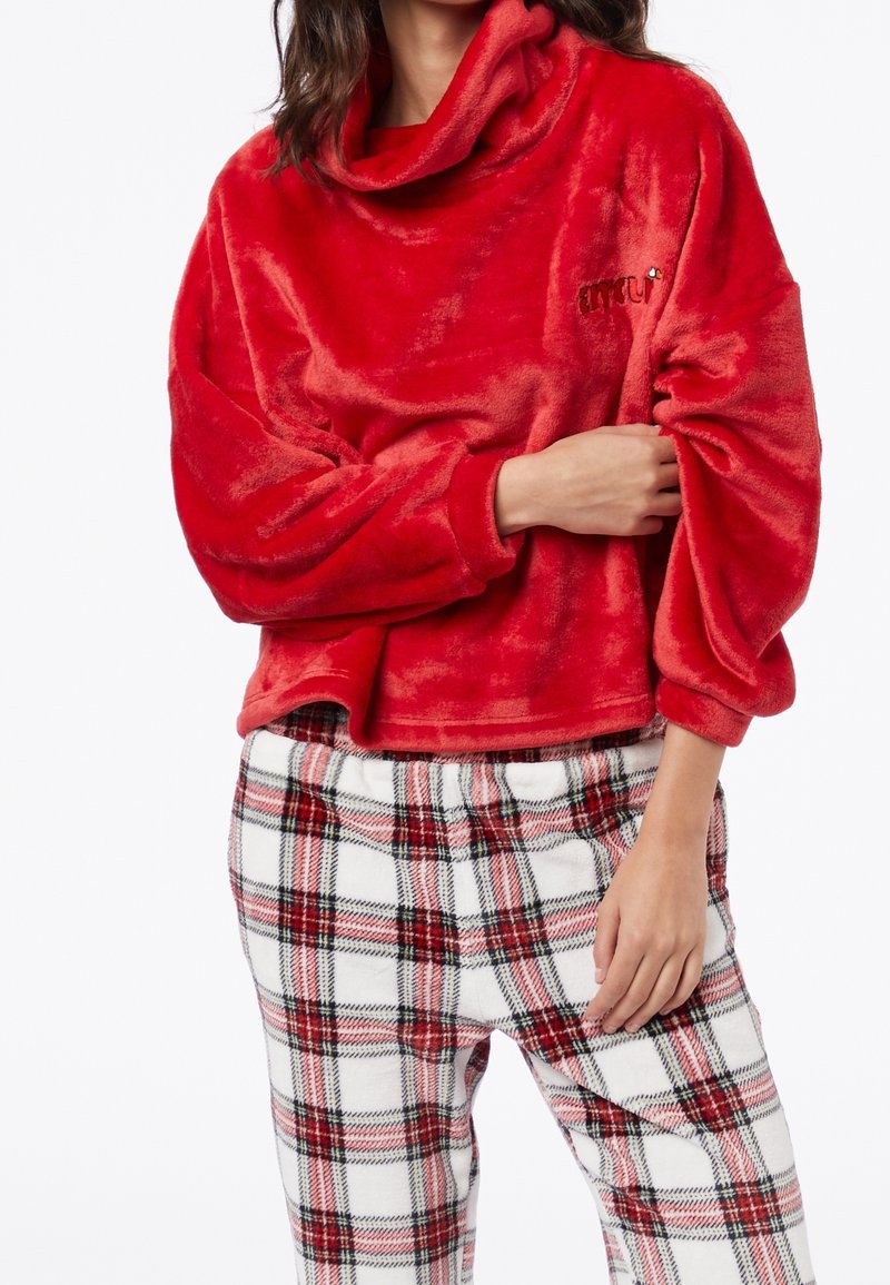 Maglione rosso in velluto con collo a cowl e maniche ampie, abbinato a pantaloni a quadri bianchi con accenti rossi e neri. Testura morbida in tutto il capo.