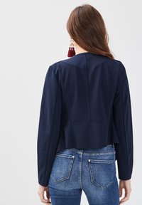 Femme aux longs cheveux bruns portant un blazer bleu marine, un jean bleu et des boucles d'oreilles à pompons rouges, debout dos à la caméra.