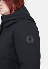 Giacca nera imbottita con collo alto e cappuccio, con una toppa rotonda con logo nero sul braccio sinistro. Tessuto dalla texture liscia.