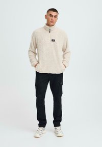 Cremfarbener Fleecepullover mit strukturiertem Oberflächen, hohem Kragen und Reißverschluss, kombiniert mit schwarzen Cargo-Hosen, die Seitentaschen und tapered Legs aufweisen.