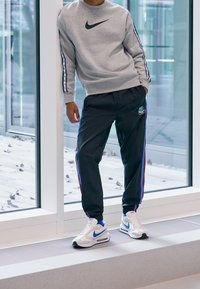 Person som bär en grå Nike-tröja, svarta Nike-träningsbyxor med sidostripes, och vita Nike-sneakers står vid ett stort fönster.