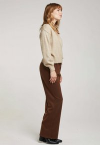 Pull en maille beige avec un col en V et des poignets côtelés, associé à un pantalon large marron. Des bottes noires à talons complètent la tenue.