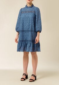 Vestido azul bordado con mangas abullonadas, cuello con volantes, intrincados patrones recortados y un dobladillo acampanado; combinado con tacones negros con tiras.