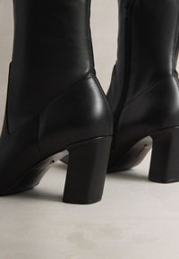 Svarta ankelstövlar i läder med klumpig klack, som har en slät textur och en sidodragkedja. Angularisk design med subtila sömnadsdetaljer.