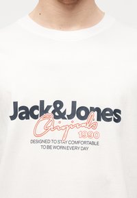 Λευκό μπλουζάκι με το μήνυμα «Jack & Jones Originals 1990» και «Designed to stay comfortable to be worn every day» τυπωμένα μπροστά με μαύρο και πορτοκαλί χρώμα.