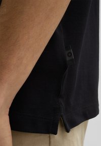 Schwarzes Poloshirt aus strukturiertem Stoff, mit seitlichen Schlitzen für mehr Flexibilität und einem kleinen Logowimpel am Saum. Der Kragen ist im traditionellen Stil.